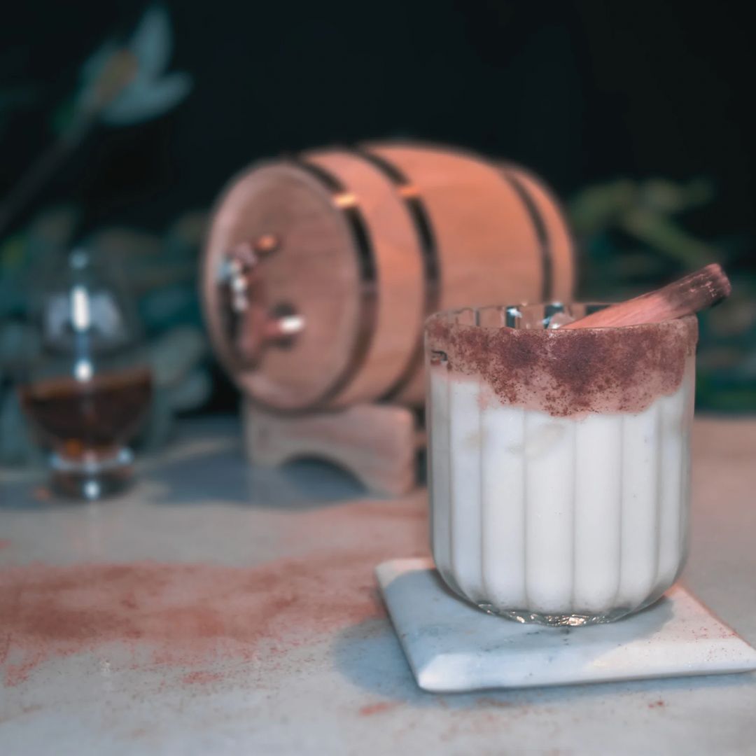 A creamy cinnamon-dusted cocktail in front of the El Padrino oak barrel