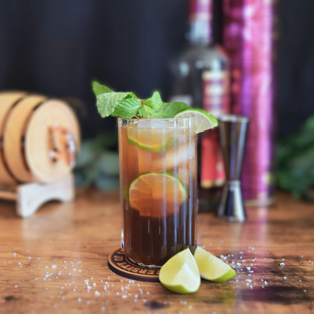 A tall cuba libre with fresh lime, poured beside the mini oak barrel